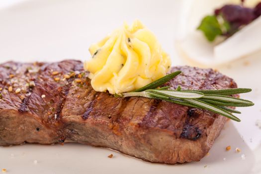 Tasty grilled beef steak topped with a twirled knob of butter and a sprig of fresh rosemary and served on a white plate, close up view