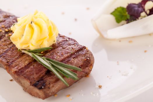 Tasty grilled beef steak topped with a twirled knob of butter and a sprig of fresh rosemary and served on a white plate, close up view