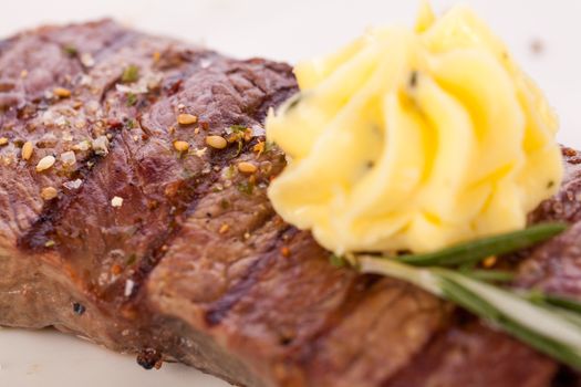 Tasty grilled beef steak topped with a twirled knob of butter and a sprig of fresh rosemary and served on a white plate, close up view