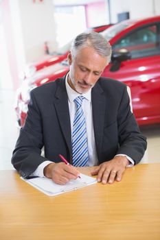 Focused salesman writing on clipboard at his desk at new car showroom