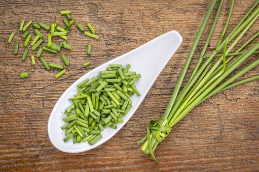 a spoon of chopped fresh chives with a bunch against rustic  wood, top view