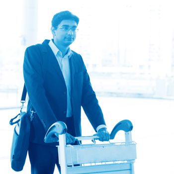 Asian Indian businessman walking with airport trolley, in blue tone.