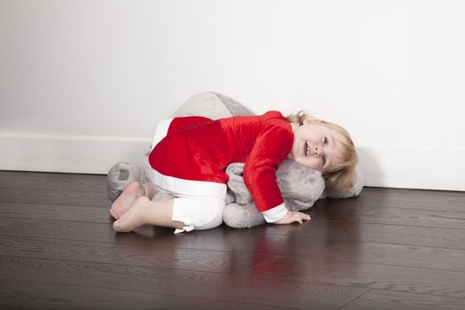 one year age caucasian blonde cute lovely tender baby barefoot Santa Claus Christmas holidays disguise lying embraced over grey plush doll on brown wooden floor white wall