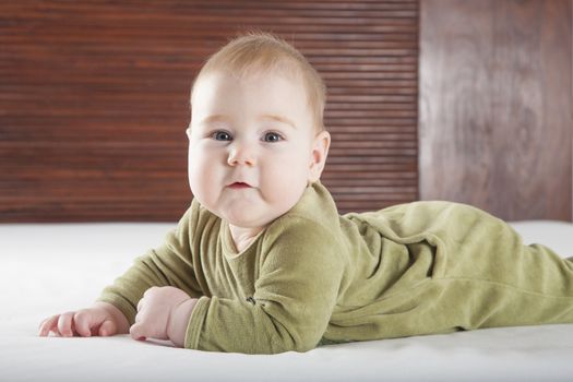 six months age blonde baby green velvet onesie lying on white sheet bed with brown wood background smiling happy face