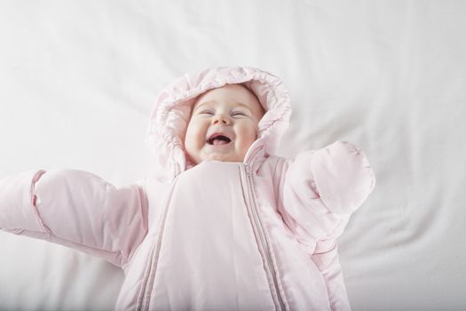 six months age cute baby dressed up in pink fluffy winter snowsuit snug hoodie clothes lying on white sheet bed smiling happy face