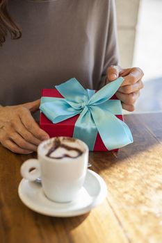 white small cup cappuccino coffee with chocolate heart and woman hands open red and blue gift box on light brown wooden table
