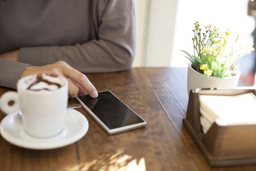 woman hand touch phone next to white cup cappuccino coffee with chocolate heart on light brown wooden table