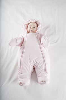 six months age cute baby dressed up in pink fluffy winter snowsuit snug hoodie clothes lying on white sheet bed smiling happy face