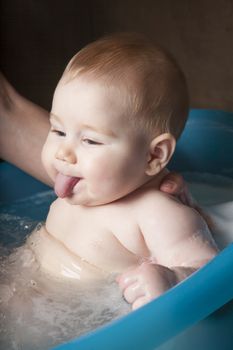 six months age blonde baby body and face sticking out tongue washing by woman mother hands in blue little plastic bath indoor with brown background