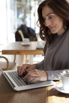 brunette mauve sweater woman typing keyboard pc laptop with white small cup cappuccino coffee on light brown wooden table cafe