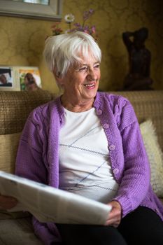 Senior woman reading morning newspaper, sitting in her favorite chair in her living room, looking happy