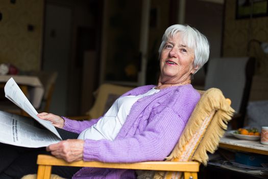 Senior woman reading morning newspaper, sitting in her favorite chair in her living room, looking happy