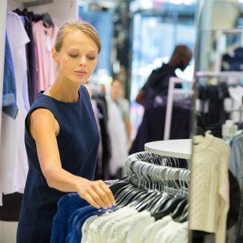 Beautiful young female shopper in a clothing store, chossing a blouse/t-shirt/garments (shallow DOF; color toned image)