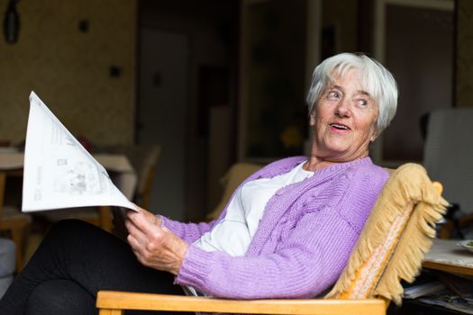 Senior woman reading morning newspaper, sitting in her favorite chair in her living room, looking happy