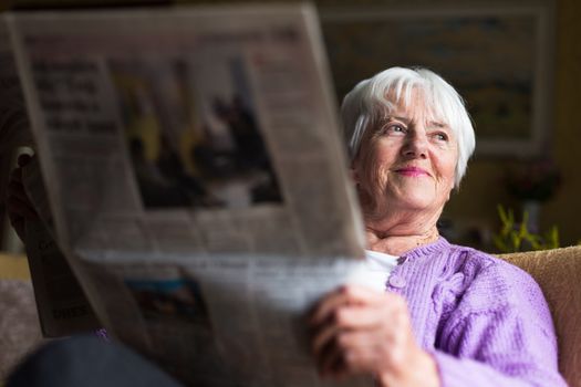 Senior woman reading morning newspaper, sitting in her favorite chair in her living room, looking happy