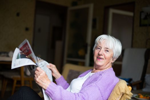 Senior woman reading morning newspaper, sitting in her favorite chair in her living room, looking happy