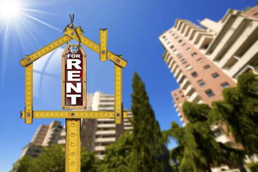 Yellow wooden meter ruler in the shape of house and label with text for rent. For rent real estate sign with tall and blurred buildings in the background