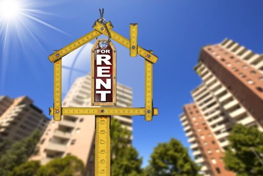 Yellow wooden meter ruler in the shape of house and label with text for rent. For rent real estate sign with tall and blurred buildings in the background