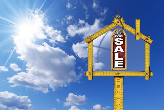 Yellow wooden meter ruler in the shape of house and label with text for sale. For sale real estate sign on blue sky with clouds and sun rays 