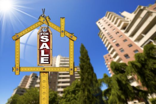 Yellow wooden meter ruler in the shape of house and label with text for sale. For sale real estate sign with tall and blurred buildings in the background
