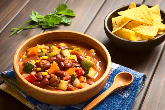 Wooden bowl of vegetarian chili dish made with kidney bean, carrot, zucchini, bell pepper, sweet corn, tomato, onion, garlic, with tortilla chips in the back, photographed with natural light (Selective Focus, Focus in the middle of the dish)   