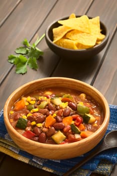 Wooden bowl of vegetarian chili dish made with kidney bean, carrot, zucchini, bell pepper, sweet corn, tomato, onion, garlic, with tortilla chips and fresh coriander in the back, photographed with natural light (Selective Focus, Focus in the middle of the dish)   
