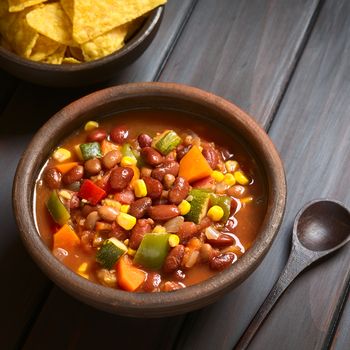 Rustic bowl of vegetarian chili dish made with kidney bean, carrot, zucchini, bell pepper, sweet corn, tomato, onion, garlic, with tortilla chips above, photographed with natural light (Selective Focus, Focus in the middle of the dish)   