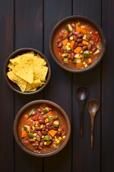 Overhead shot of two rustic bowls of vegetarian chili dish made with kidney bean, carrot, zucchini, bell pepper, sweet corn, tomato, onion, garlic, with tortilla chips on the side, photographed on dark wood with natural light