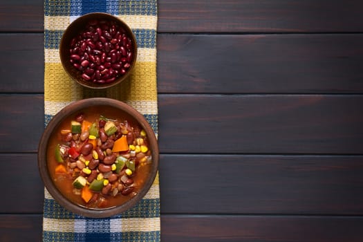 Overhead shot of a rustic bowl of vegetarian chili dish made with kidney bean, carrot, zucchini, bell pepper, sweet corn, tomato, onion, garlic, raw kidney beans in bowl above, photographed on dark wood with natural light