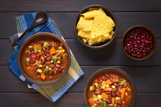 Overhead shot of two rustic bowls of vegetarian chili dish made with kidney bean, carrot, zucchini, bell pepper, sweet corn, tomato, onion, garlic, with tortilla chips and raw kidney beans on the side, photographed on dark wood with natural light