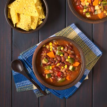 Overhead shot of rustic bowl of vegetarian chili dish made with kidney bean, carrot, zucchini, bell pepper, sweet corn, tomato, onion, garlic, with tortilla chips on the side, photographed on dark wood with natural light