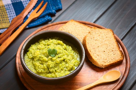 Rustic bowl of homemade zucchini and parsley spread garnished with fresh parsley leaf, slices of wholegrain bread on the side, photographed on dark wood with natural light (Selective Focus, Focus on the leaf) 