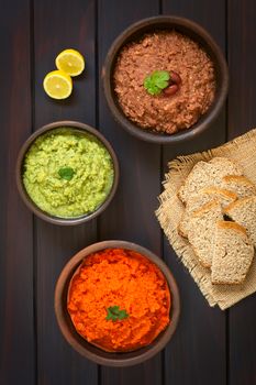 Overhead shot of rustic bowls of homemade vegetable spreads (red kidney bean, zucchini and parsley, carrot and red bell pepper), slices of wholegrain bread and lemon on the side, photographed on dark wood with natural light