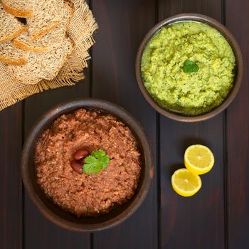 Overhead shot of rustic bowls of homemade vegetable spreads (red kidney bean, zucchini and parsley), slices of wholegrain bread and lemon on the side, photographed on dark wood with natural light