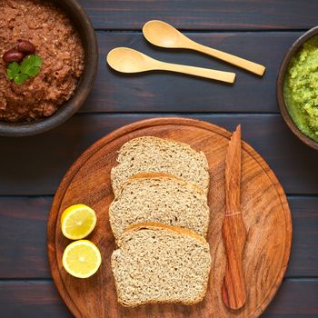 Overhead shot of wholegrain bread slices on wooden plate with two rustic bowls of homemade vegetable spreads (red kidney bean, zucchini and parsley), photographed on dark wood with natural light