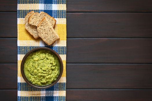 Overhead shot of rustic bowl of homemade zucchini and parsley spread with slices of wholegrain bread on kitchen towel, photographed on dark wood with natural light