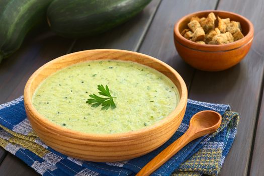 Cream of zucchini soup in wooden bowl garnished with parsley leaf, croutons in the back, photographed with natural light (Selective Focus, Focus on the parsley leaf)