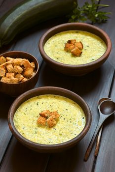 Two rustic bowls of cream of zucchini soup with homemade croutons on top, photographed on dark wood with natural light (Selective Focus, Focus on the croutons on the first soup)