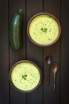 Overhead shot of two rustic bowls of cream of zucchini soup garnished with parsley leaf, photographed on dark wood with natural light
