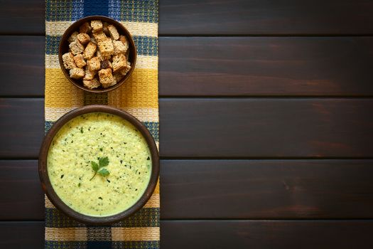 Overhead shot of a rustic bowl of cream of zucchini soup garnished with parsley leaf, with a bowl of homemade croutons on kitchen towel, photographed on dark wood with natural light