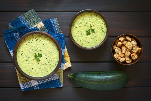 Overhead shot of two rustic bowls of cream of zucchini soup garnished with parsley leaf, with a bowl of homemade croutons and a raw zucchini on the side, photographed on dark wood with natural light