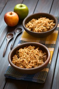 Two rustic bowls of baked apple crumble or crisp on kitchen towel, wooden spoons and fresh apples on the side, photographed on dark wood with natural light (Selective Focus, Focus one third into the first crumble)