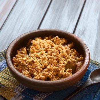 Rustic bowl of baked apple crumble or crisp on kitchen towel, wooden spoon on the side, photographed on dark wood with natural light (Selective Focus, Focus one third into the first crumble)