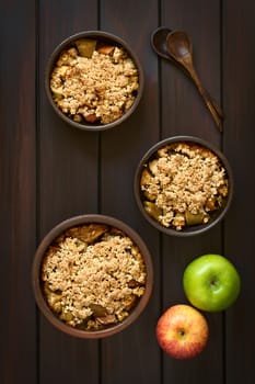 Overhead shot of three rustic bowls of baked apple crumble or crisp, wooden spoons and fresh apples on the side, photographed on dark wood with natural light