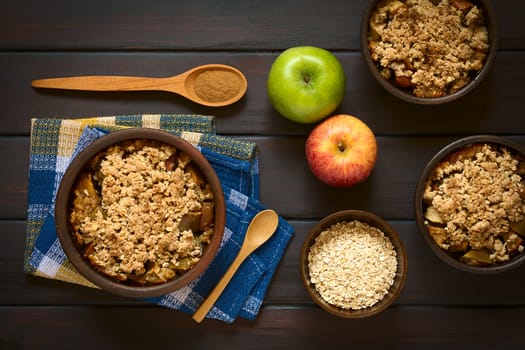 Overhead shot of three rustic bowls of baked apple crumble or crisp, spoonful of cinnamon powder, fresh apples and raw rolled oats on the side, photographed on dark wood with natural light