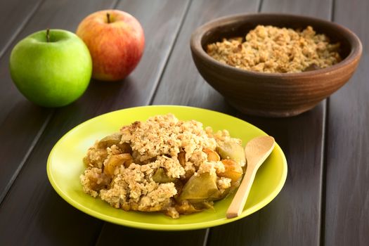 Freshly baked apple crumble or crisp served on plate with wooden spoon, fresh apples and a rustic bowl of apple crumble in the back, photographed on dark wood with natural light (Selective Focus, Focus one third into the crumble)