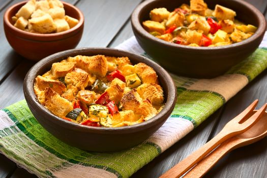 Savory baked vegetarian bread pudding made of zucchini, bell pepper, tomato and diced baguette, seasoned with thyme and parsley in rustic bowl on kitchen towel, photographed with natural light (Selective Focus, Focus on the zucchini pieces in the front)