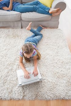 High angle view of girl using laptop while lying on rug with mother relaxing on sofa at home