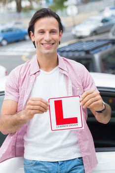 Learner driver smiling and holding l plate in front of his car 