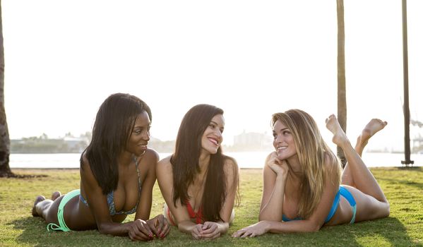 Three women lying on their belly, on the grass in a park. Backlit, looking at each other.